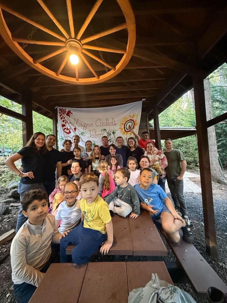 A large group of adults and children gathered under a wooden pavilion, smiling and posing for a photo in front of a banner that reads 'Magyar Családi Tábor.'