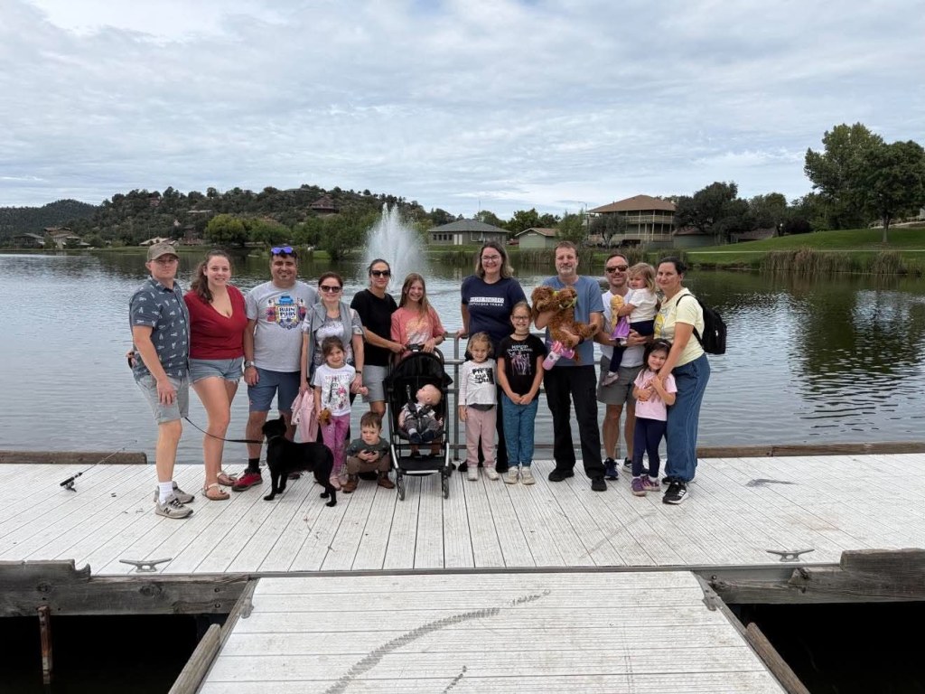 A group of people, including adults and children, gathered on a dock by a lake with a fountain in the background. Some individuals are holding dogs and children, while others are enjoying the outdoors.
