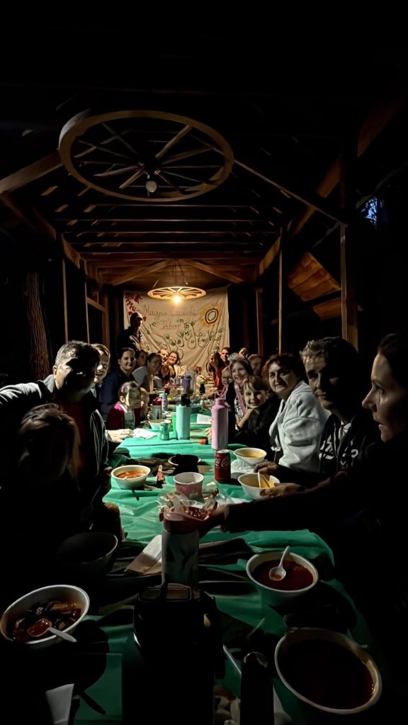 A long table filled with bowls of food, soft drinks, and utensils, surrounded by a group of people smiling and enjoying a gathering in a dimly-lit space.