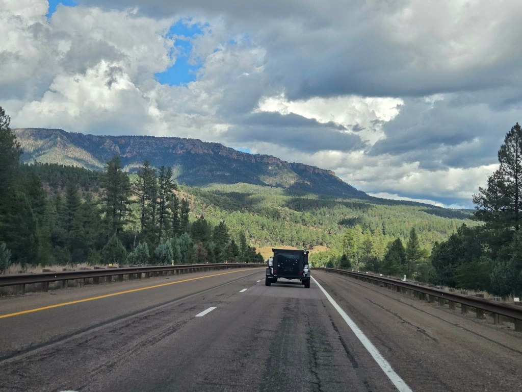 A black vehicle driving on a two-lane road surrounded by green pine trees and mountains under a partly cloudy sky.
