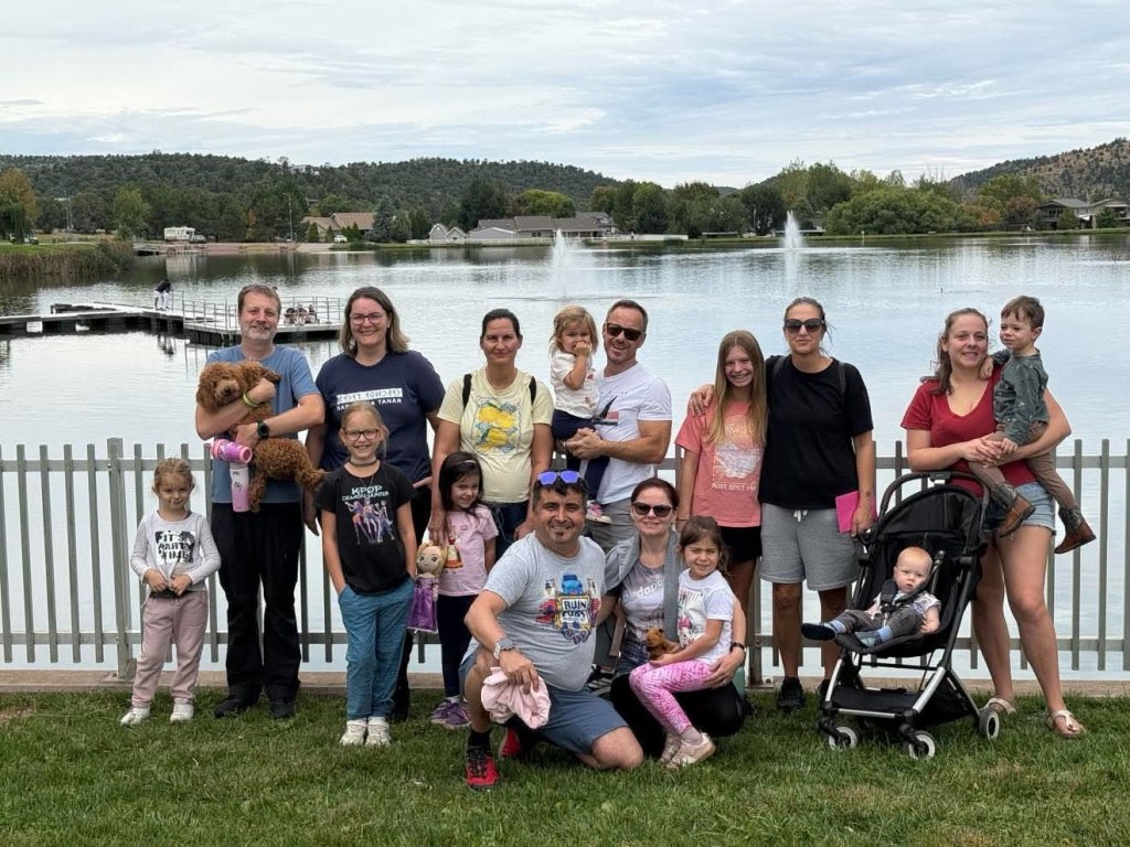 Group photo of a family gathering by a lake, with children and adults holding toys and pets, and a stroller in the foreground.